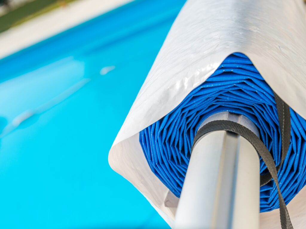 Close-up of a rolled-up blue pool cover attached to a metal reel beside a swimming pool, showcasing the convenience and effectiveness of modern Pool Covers.