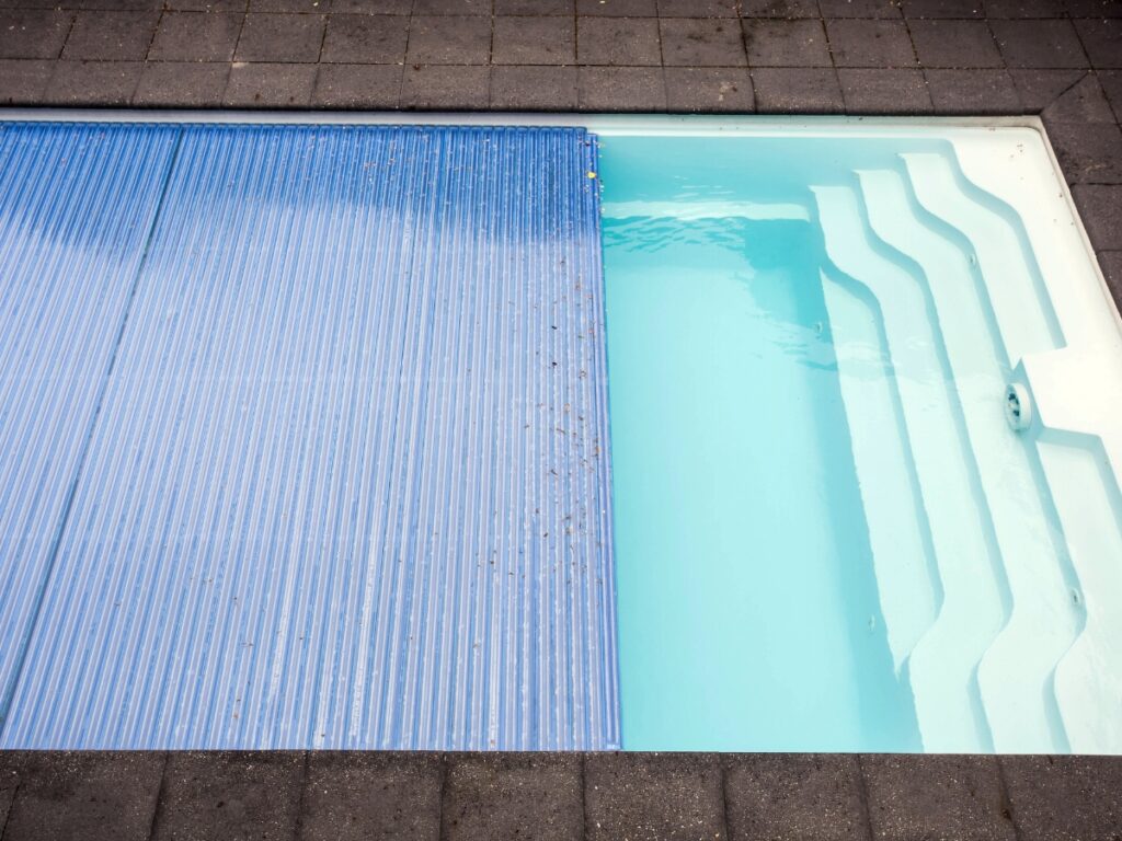 Outdoor swimming pool with white steps and a partially closed blue pool cover, surrounded by gray paving stones. Pool covers provide added safety and help keep your water clean when the pool is not in use.