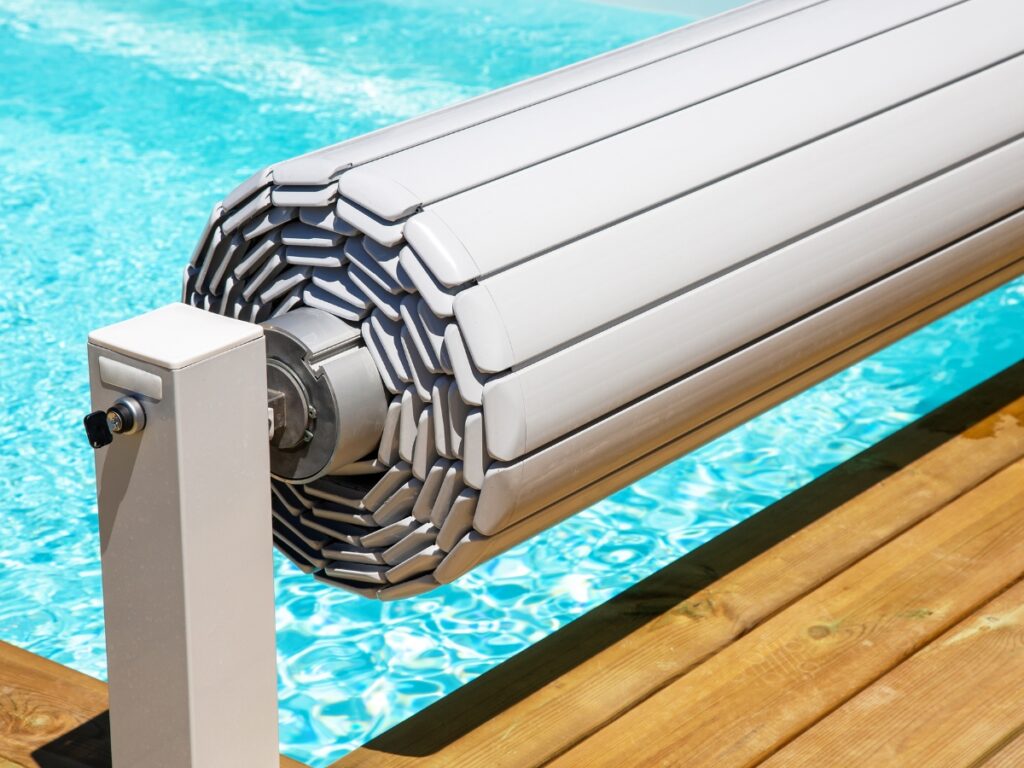 A close-up of a rolled-up pool cover on a metal reel next to a clear blue swimming pool and wooden deck highlights the practicality and convenience of Pool Covers.
