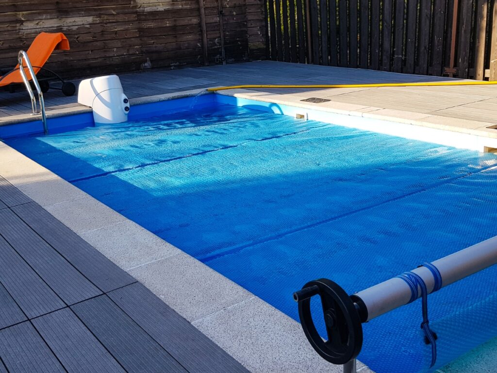 Small outdoor pool with a blue pool cover partially rolled out on a pool roller, Pool Covers equipment, a white pool skimmer, metal ladder, orange slide, and wooden fencing surrounding the area.