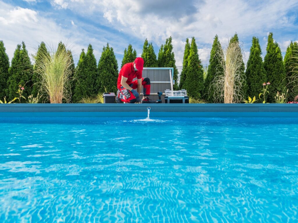 A person in red work clothes kneels by the edge of an outdoor swimming pool, appearing to perform maintenance or check the pool covers; trees and shrubs line the background.