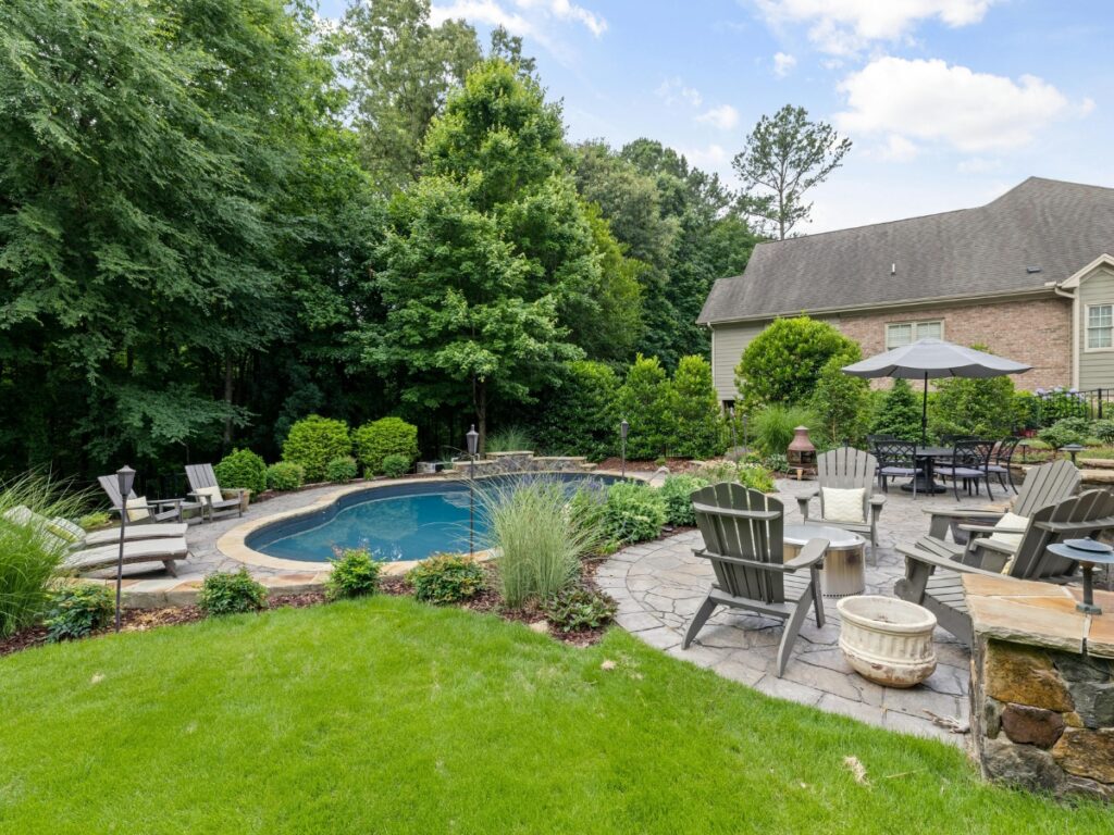 Backyard with in-ground pool, stone patio, Adirondack chairs, umbrella-covered dining area, fire pit, and lush greenery surrounding a tranquil zen garden.