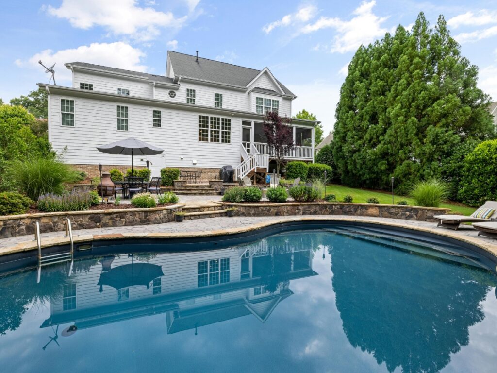 A backyard with an in-ground swimming pool, stone patio, zen garden, outdoor furniture, and landscaping, set behind a two-story white house with large windows.