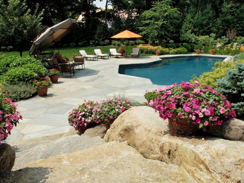 Backyard swimming pool surrounded by lounge chairs, umbrellas, stone patio, and a lush green lawn, with trees and colorful potted flowers adding a touch of zen garden tranquility in the foreground.