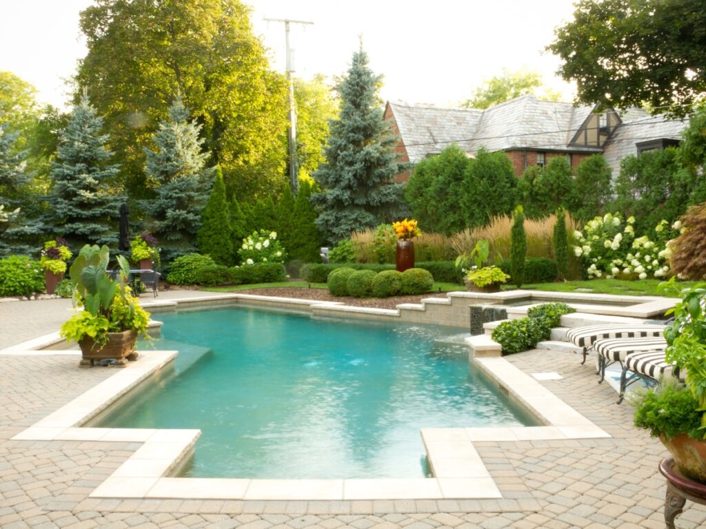 A landscaped backyard with a rectangular swimming pool, lounge chairs, potted plants, manicured greenery, and a serene zen garden, with a house in the background.