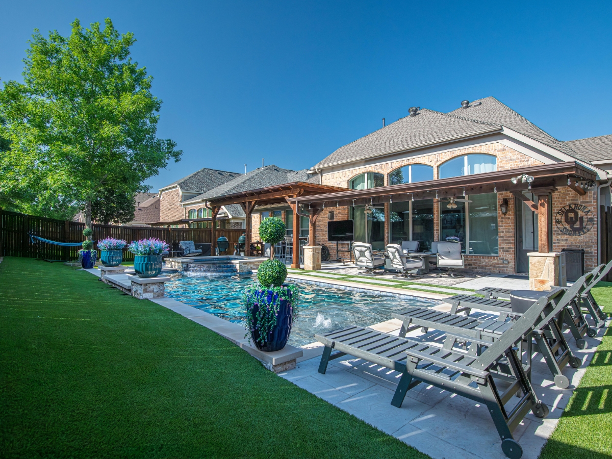 Backyard with a swimming pool, lounge chairs, potted plants, a tranquil zen garden, covered patio, and a brick house in the background under a clear blue sky.