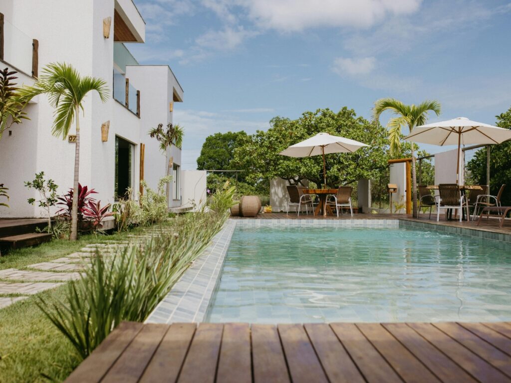 Outdoor swimming pool beside a modern white building, featuring palm trees, patio tables, chairs, umbrellas, and low maintenance landscaping under a partly cloudy sky.