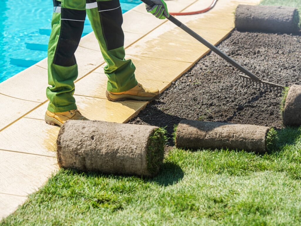 Person raking soil beside a swimming pool, preparing ground to lay rolls of turf grass for a beautiful, low maintenance landscaping solution.