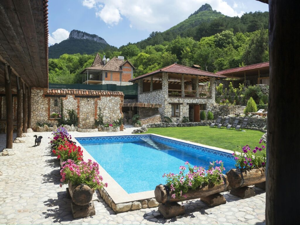 Stone courtyard with a rectangular swimming pool, surrounded by low maintenance landscaping, flowers, lounge chairs, and rustic buildings, set against a backdrop of green hills and trees.