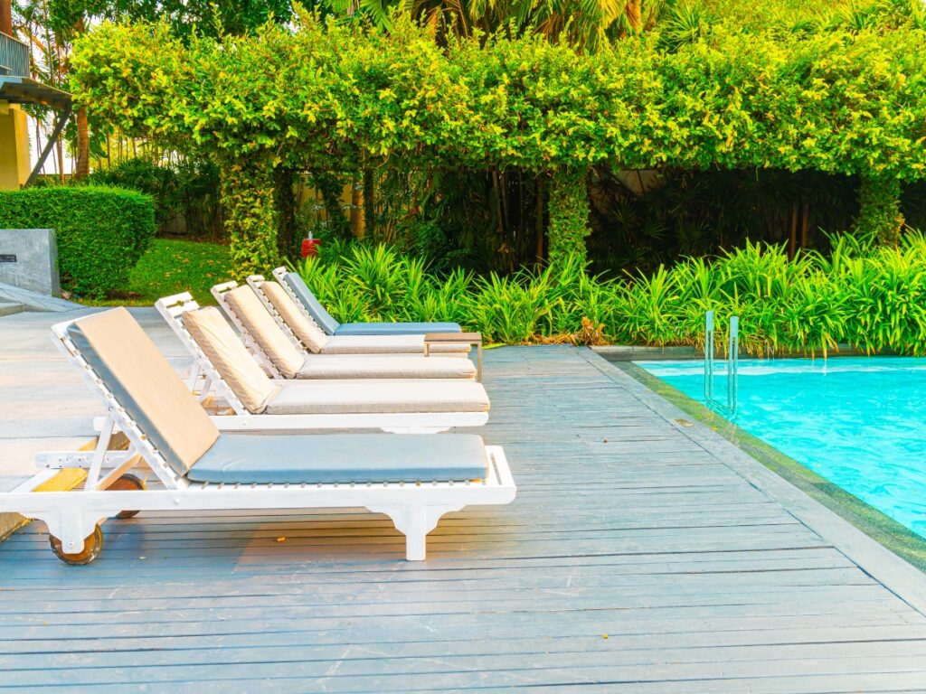 Five white lounge chairs are lined up on a wooden deck next to a swimming pool, surrounded by low maintenance landscaping with green hedges and trees in the background.