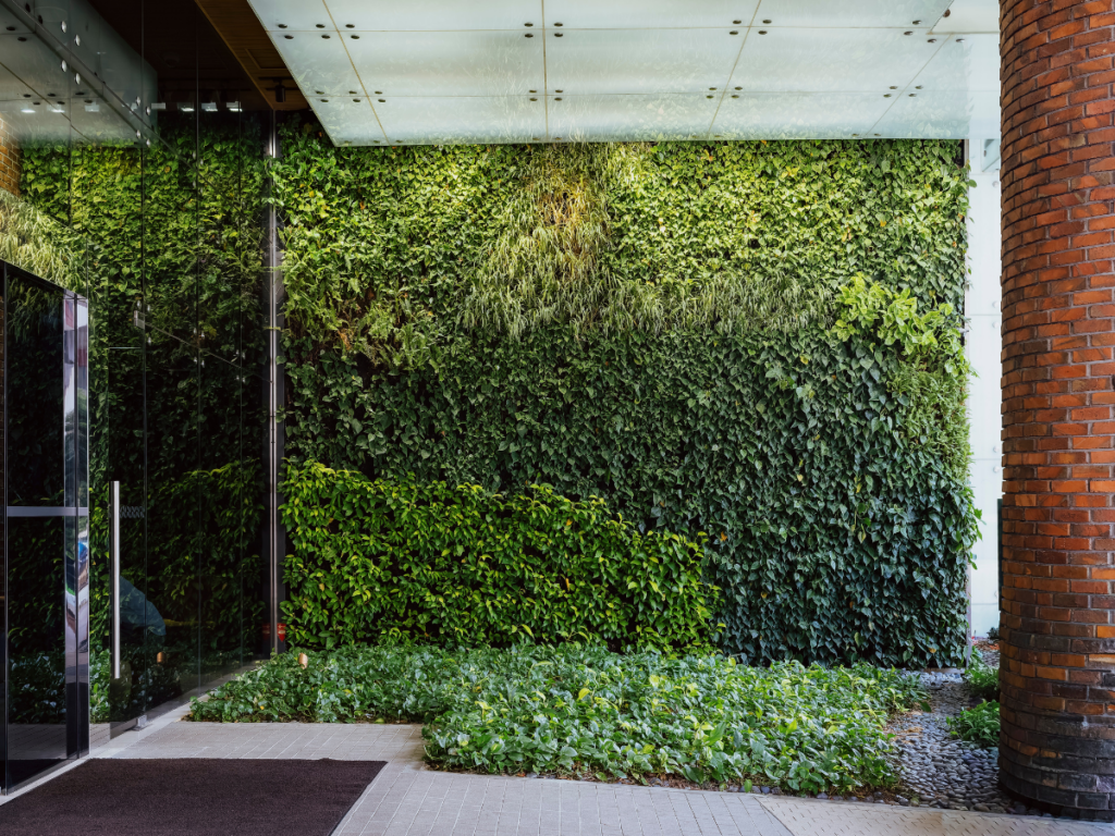 A modern building entrance features striking vertical gardens on a tall green wall, complemented by a glass canopy, brick column, and leafy ground plants beside a sleek glass door.