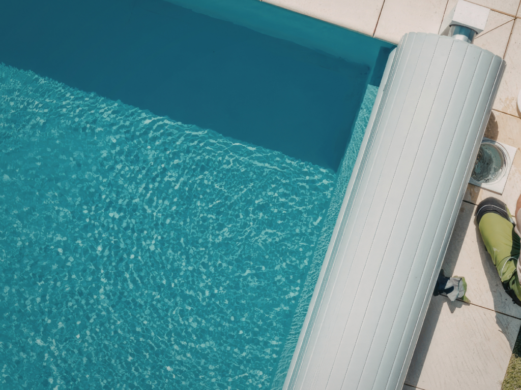 A retractable pool cover is partially rolled up beside a clean blue swimming pool, with a person's feet visible near the edge on tiled flooring—an ideal scene for your summer guide to pool season.