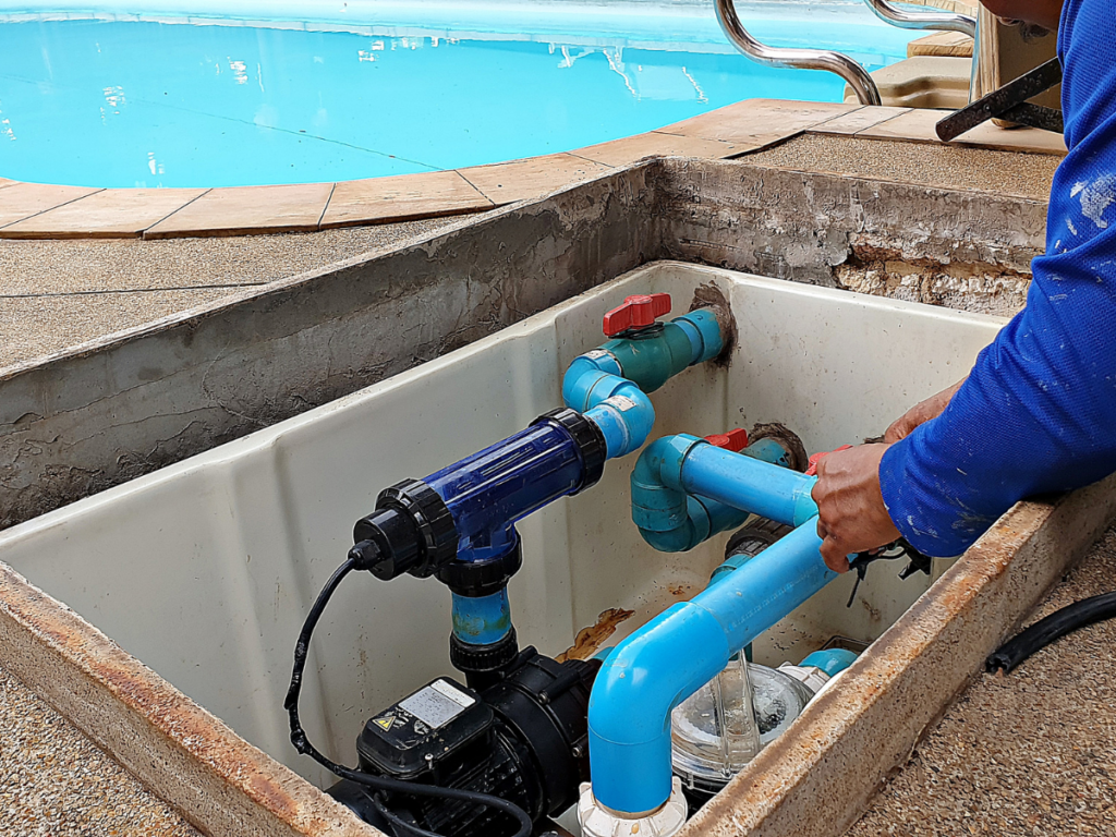 A person repairs or maintains blue PVC pool plumbing and a pump system in a concrete pit next to a swimming pool as part of their summer startup guide for pool season.