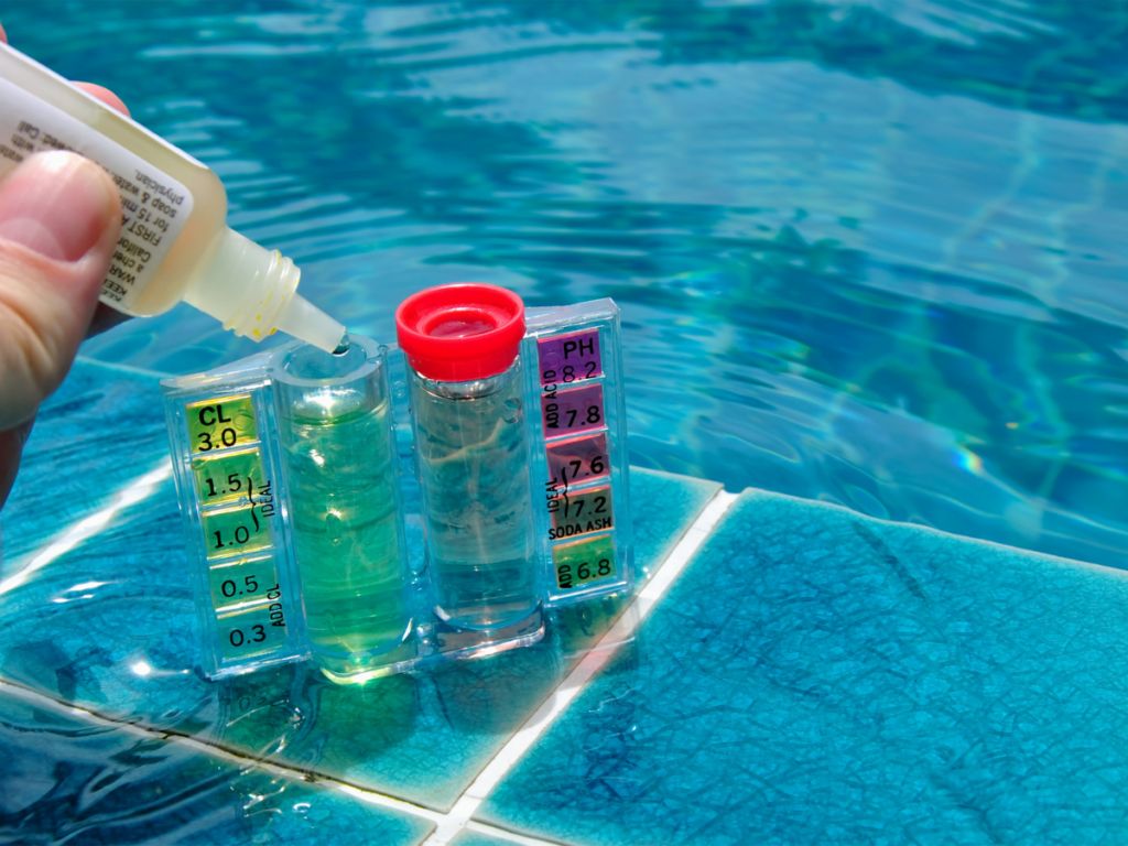 A hand adds testing solution to a pool water test kit with vials showing chlorine and pH levels by the edge of a swimming pool—an essential step in any pool season startup guide.