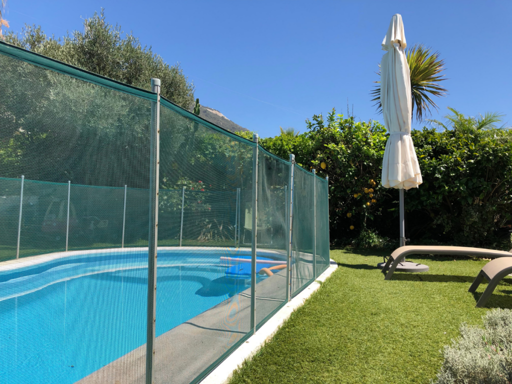 A fenced outdoor swimming pool with a pool float, surrounded by green grass, two lounge chairs, and an upright closed umbrella under a clear blue sky—perfect for diving into Pool season or following your pool startup guide this summer.