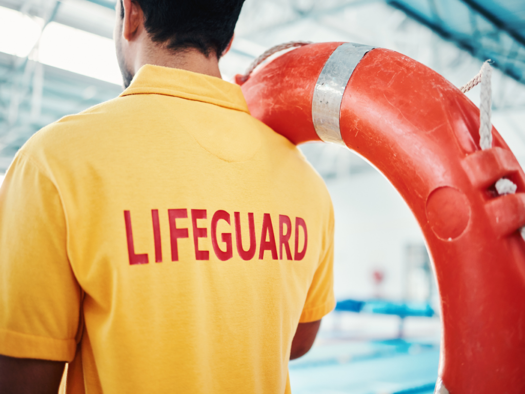 A lifeguard in a yellow shirt stands next to an orange lifebuoy at an indoor swimming pool, ready for pool season.
