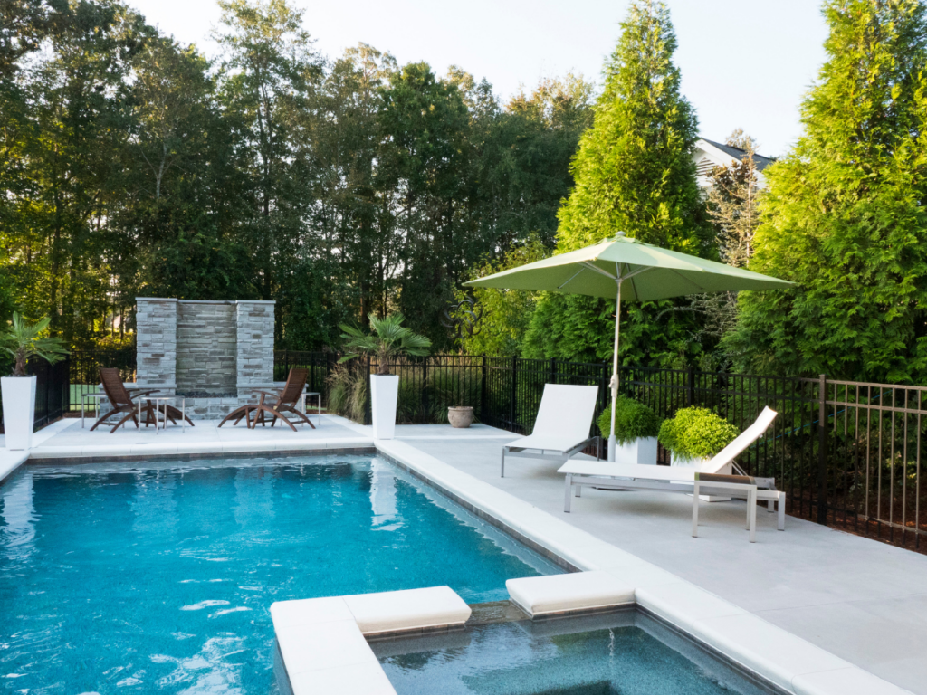 A modern outdoor pool area welcomes summer with lounge chairs, a green umbrella, a spa section, and a water feature—all surrounded by trees and a black metal fence, perfect for enjoying pool season.