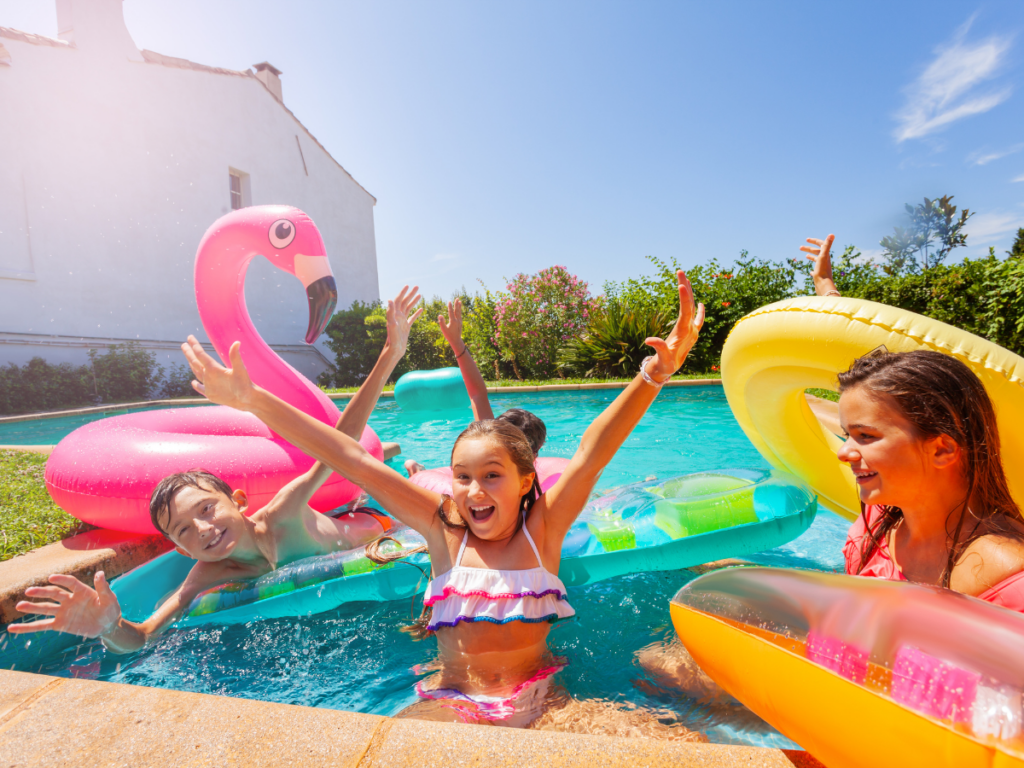 Four children in swimsuits play and smile on colorful inflatable pool floats, including a pink flamingo, enjoying pool games in an outdoor swimming pool on a sunny day.