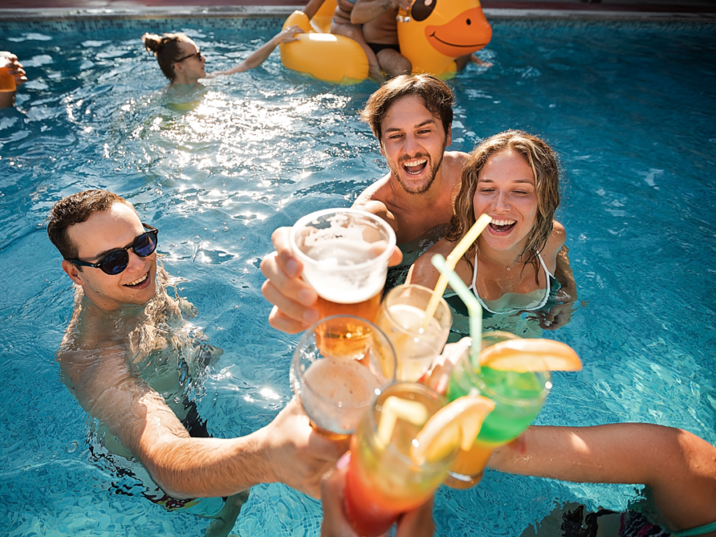 A group of people in a swimming pool raise colorful drinks together, smiling and celebrating a sunny day after enjoying exciting pool games.