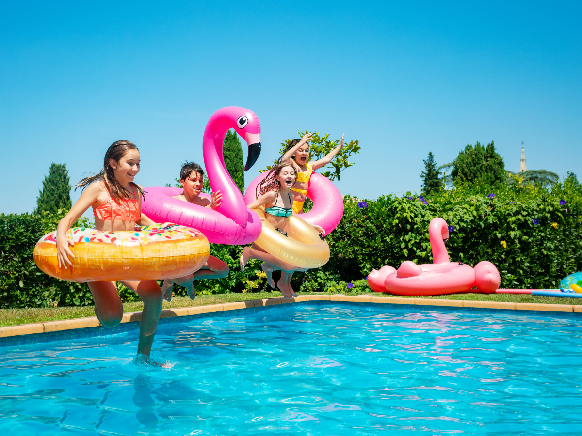Four children with inflatable pool floats jump into an outdoor swimming pool on a sunny day, ready for exciting pool games, with greenery and blue sky in the background.