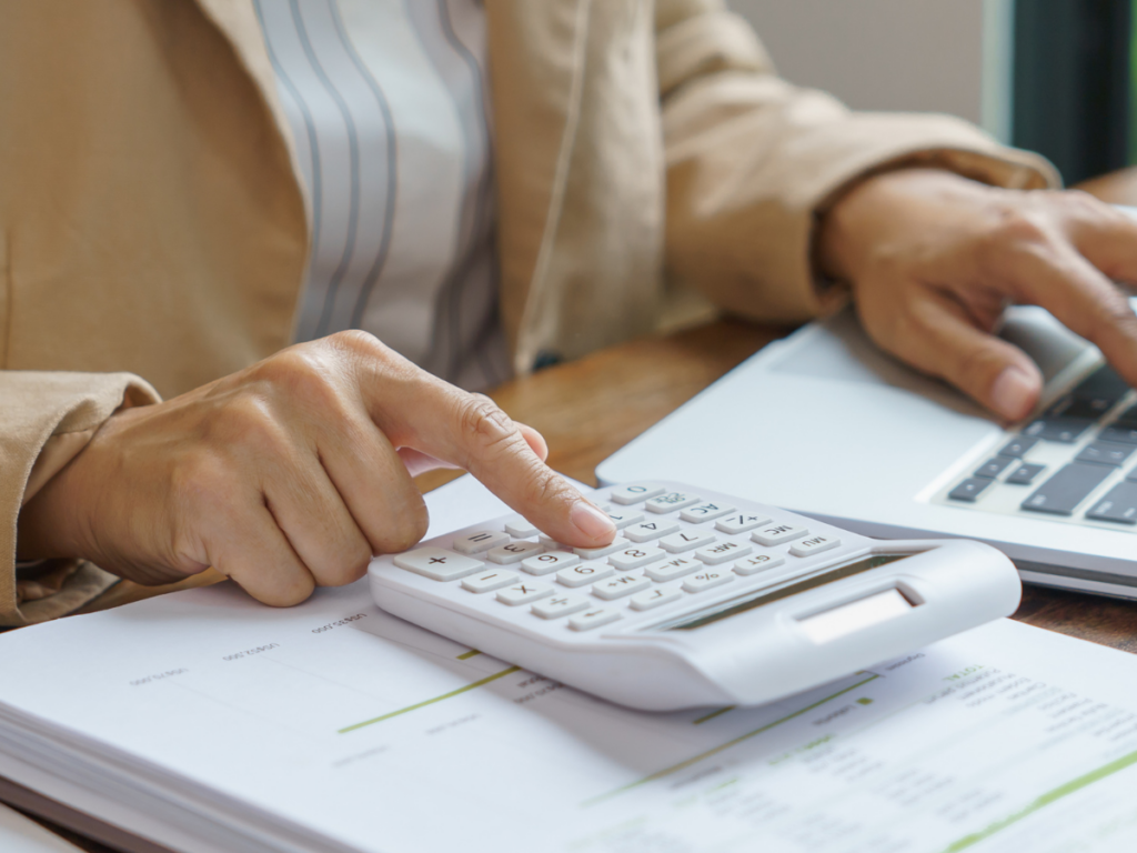 Person using a calculator and typing on a laptop, with documents spread on a wooden desk, researching the latest trends in beach entry pools.