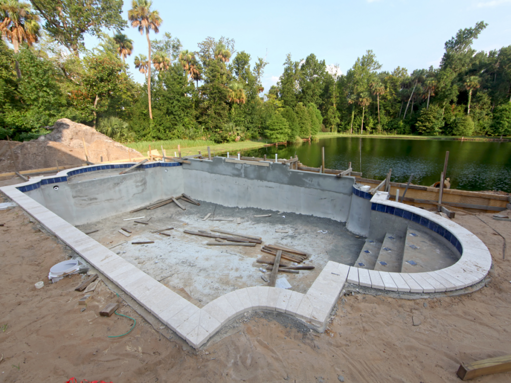 An empty swimming pool under construction, featuring a beach entry pool design, is surrounded by sand and wooden planks, with a lake and trees in the background.