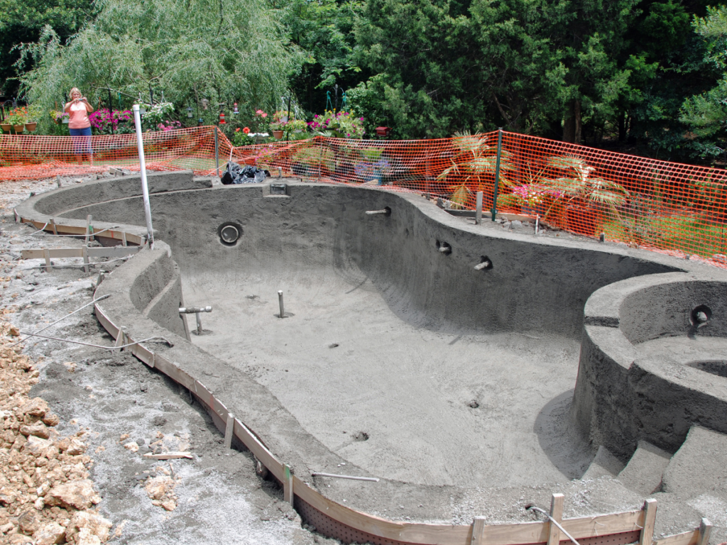 Partially constructed in-ground pool with exposed concrete walls, surrounded by orange safety fencing; a person stands near garden plants in the background, perhaps considering the fiberglass pool benefits for a smoother installation and enhanced durability.