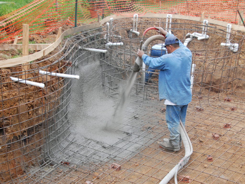 A worker sprays concrete onto rebar framework during the construction of an in-ground swimming pool, a traditional method that contrasts with fiberglass pool benefits such as faster installation and lower long-term maintenance.