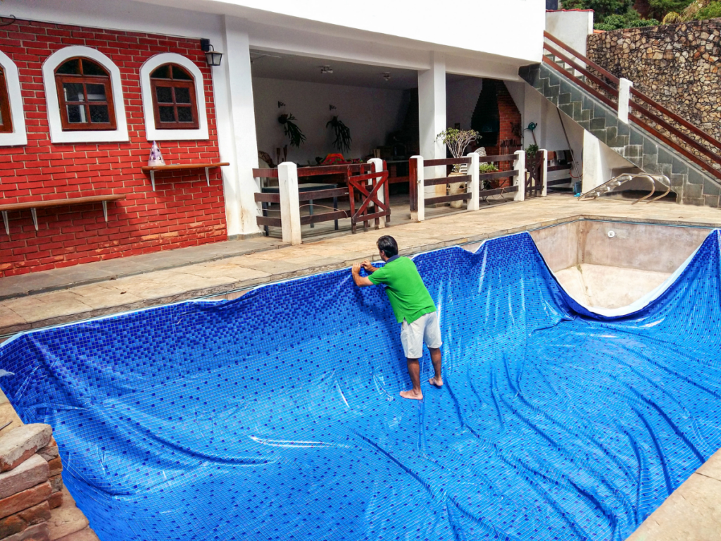 A person in a green shirt installs a blue pool liner in an empty swimming pool beside a building with red brick walls and outdoor seating, highlighting some of the fiberglass pool benefits such as durability and easy maintenance.