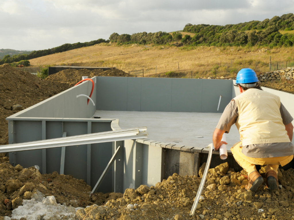 A construction worker in a blue helmet inspects the early stages of an outdoor swimming pool build at a rural site, highlighting the fiberglass pool benefits such as durability and easy installation.