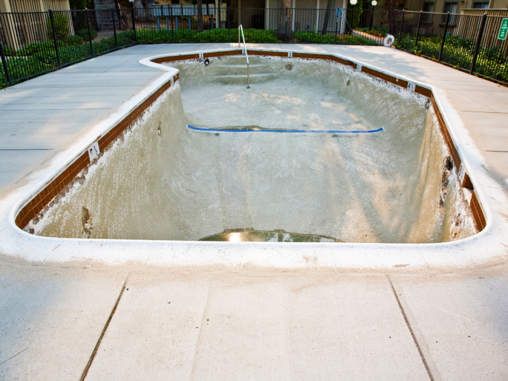 Empty outdoor swimming pool with dirty, rough surface and visible cleaning tools, surrounded by a fence and concrete pavement, in a residential or hotel setting during daylight. The neglected appearance highlights the low maintenance and durability advantages among fiberglass pool benefits over traditional pools like this one.