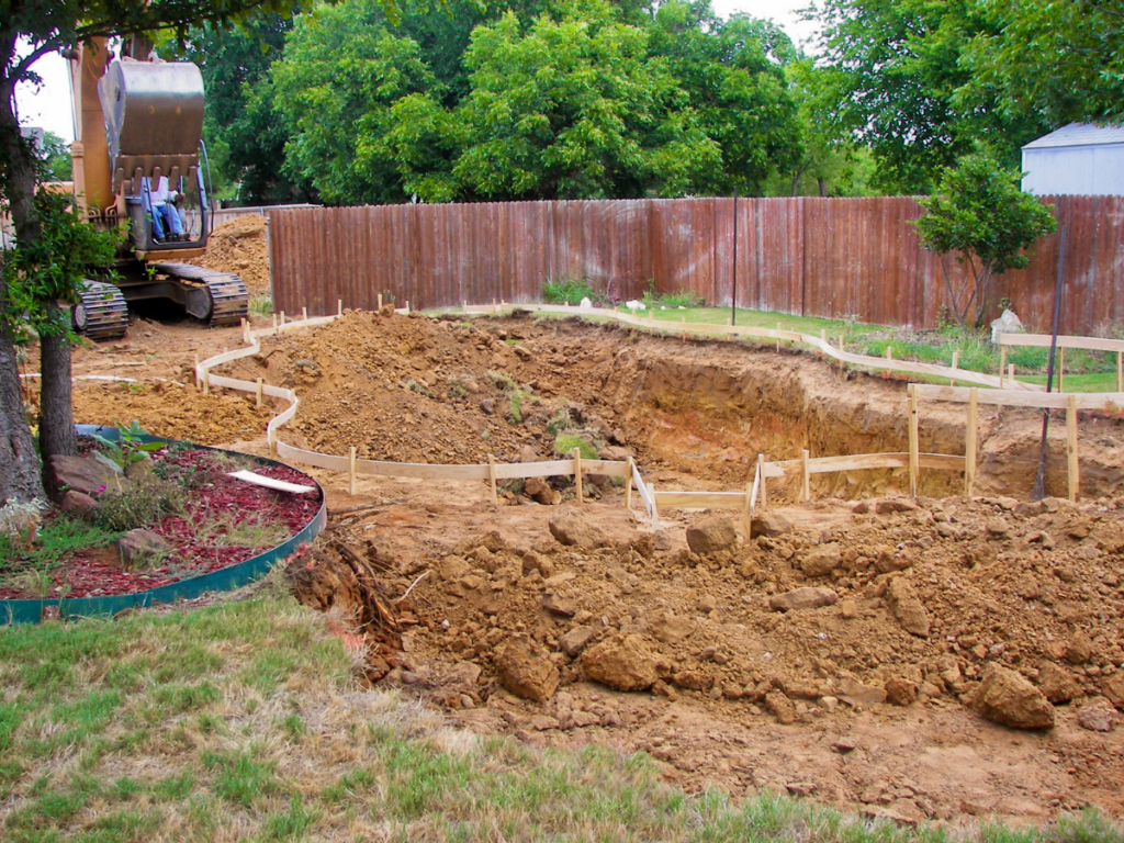 An excavator is digging a large hole in a backyard, with wooden forms outlining the shape of a future in-ground pool and piles of dirt surrounding the site. Homeowners considering this project may be drawn to fiberglass pool benefits, such as quick installation and low maintenance, making them an appealing choice for backyard transformations.