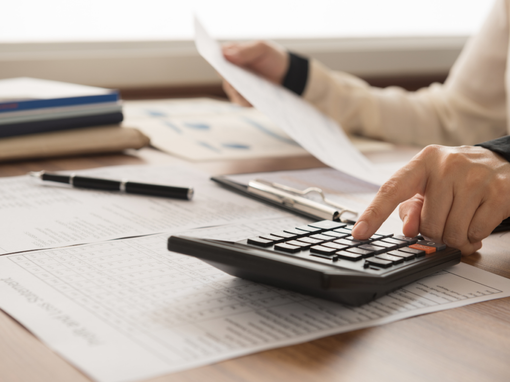 Person using a calculator at a desk, holding a document, with financial papers, a pen, and folders nearby&mdash;perhaps carefully budgeting for home improvements or researching plunge pool ideas.
