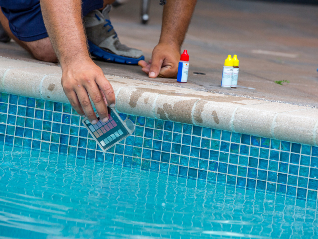 Person testing swimming pool water with a test kit near the pool edge; two bottles of testing chemicals are placed on the tiled edge, an important step not only for water balance but also for effective pool stain removal.