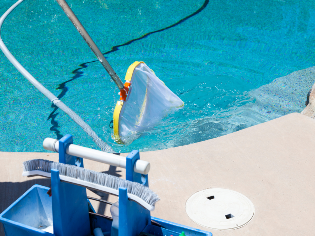 A pool skimmer net is being used to clean debris from a swimming pool. A cleaning caddy with brushes and pool stain removal essentials is nearby on the pool deck.