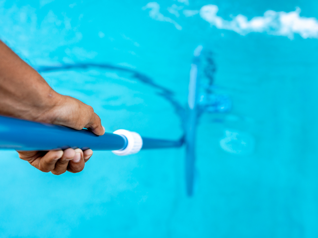 A hand holding a blue pool cleaning pole with a brush, submerged in clear blue pool water, demonstrates effective pool stain removal.