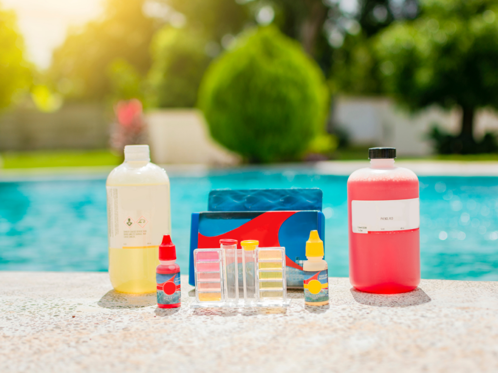 Bottles of pool chemicals, including solutions for pool stain removal, and a pool water testing kit are arranged on a stone surface beside a swimming pool on a sunny day.