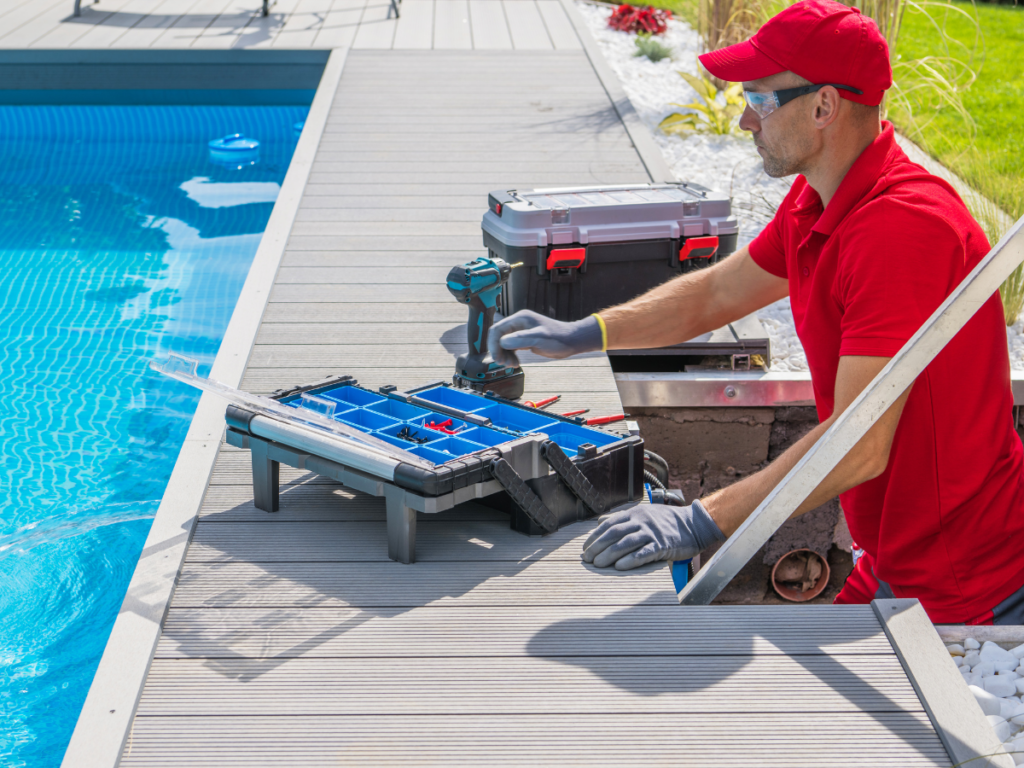 A maintenance worker in red uniform and gloves repairs pool equipment beside an outdoor pool, with tools and a toolbox on the deck, ensuring thorough pool stain removal as part of regular upkeep.