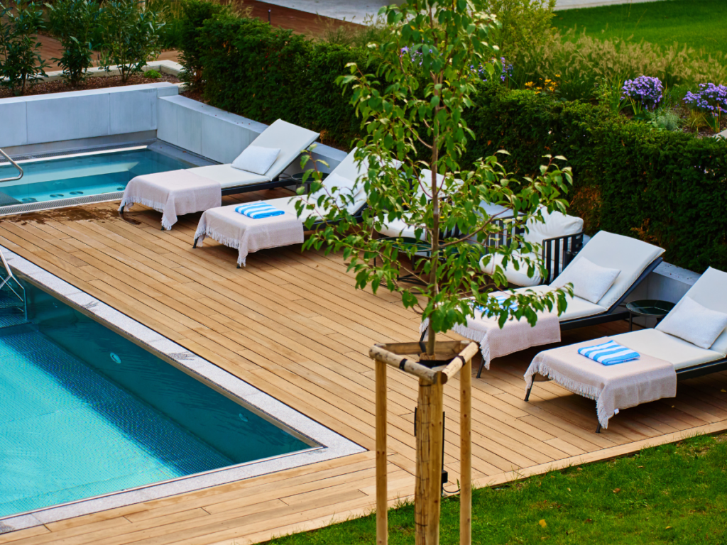 Outdoor pool area with wooden deck crafted from some of the best pool deck materials, four lounge chairs with white towels, a small tree, green grass, hedges, and flowering plants.