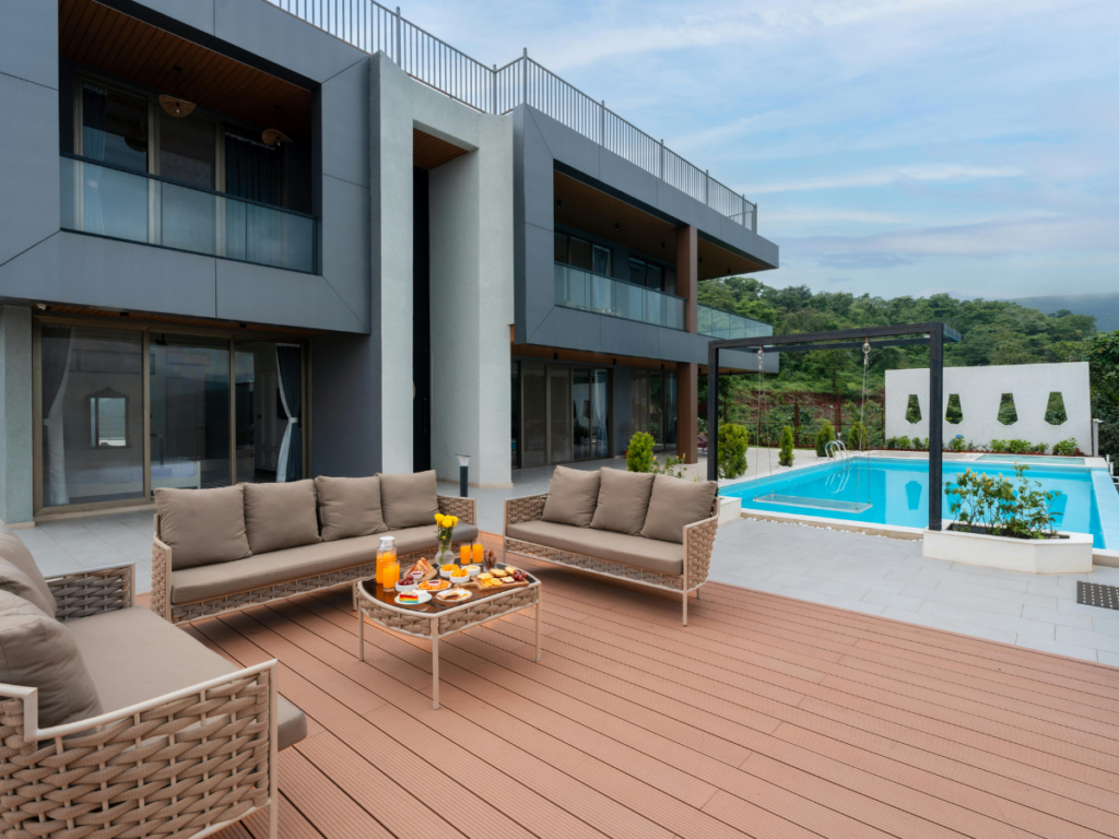 Modern patio with brown outdoor sofas, table set with drinks and snacks, and a swimming pool beside a contemporary two-story house featuring the best pool deck materials. Trees and hills are visible in the background.